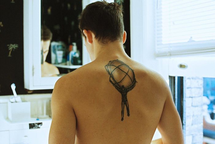 Man with a back tattoo showing an Atlas figure holding a globe, standing in a bathroom.