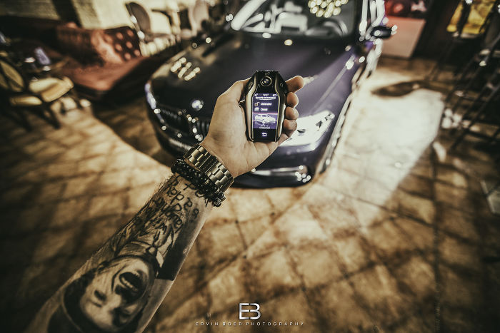 Person with arm tattoos holding a car key fob, luxury car in background.