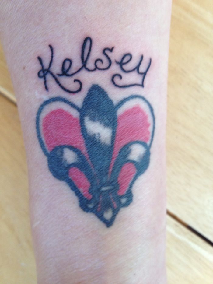 Tattoo of a red and black fleur-de-lis with cursive text above on a forearm.