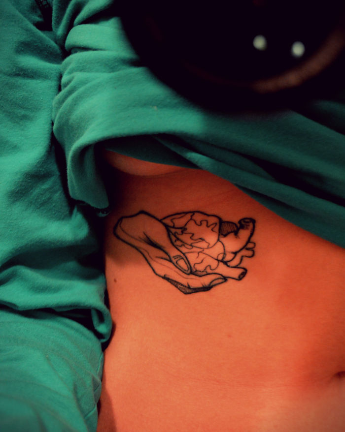 Tattoo of a hand holding an anatomical heart on the side, partially covered by a green shirt.