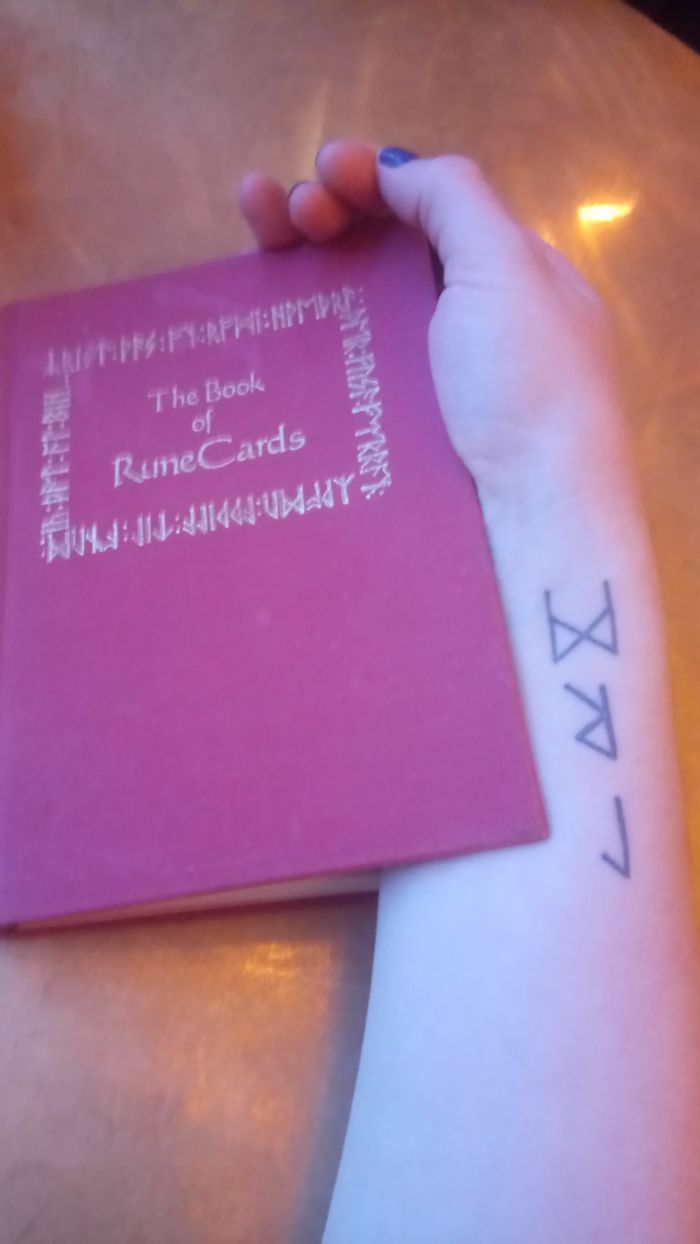 A tattoo of geometric symbols on an arm, holding "The Book of RuneCards" on a wooden table.