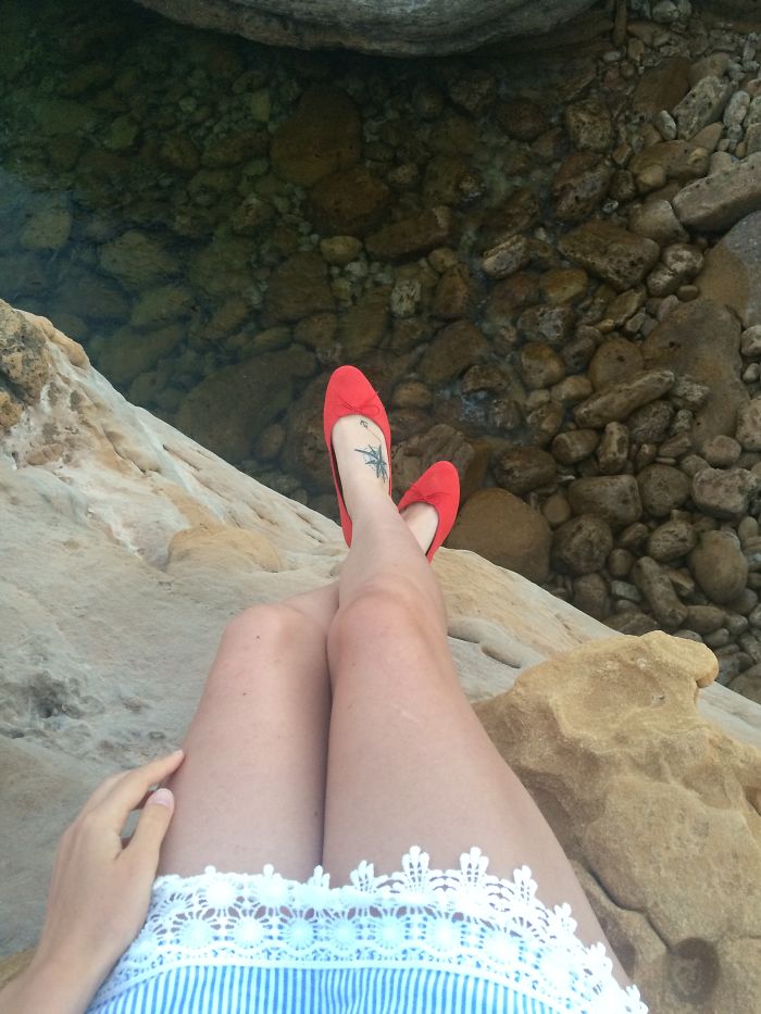 A person sitting on rocks near water with a tattoo on their leg and wearing red shoes.