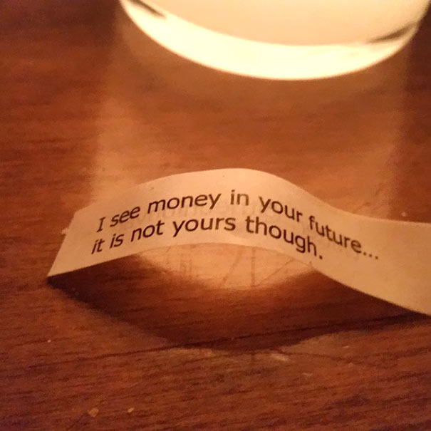 Sassy fortune cookie saying about seeing money in the future but not yours, on a wooden table near a lit candle.