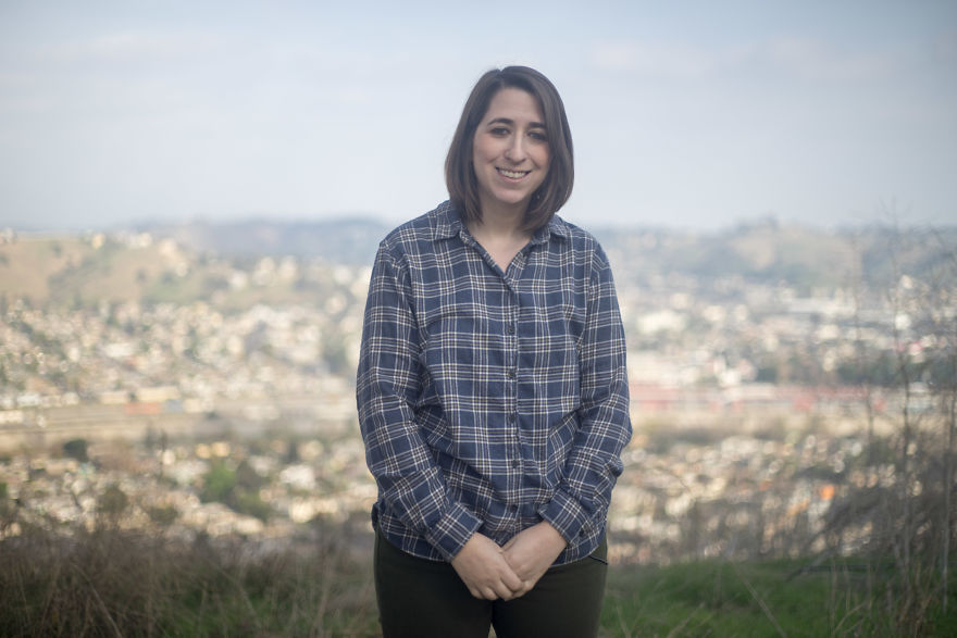 Young woman standing outdoors, smiling and wearing a plaid shirt, related to interviewing people on their birthdays.