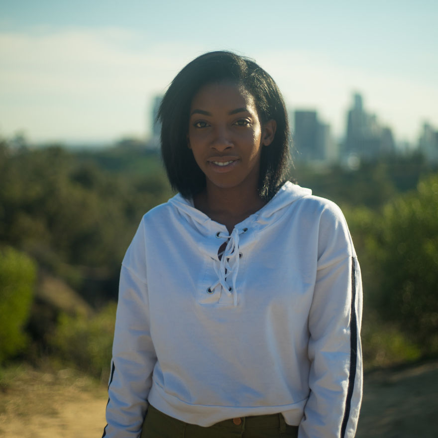 Young woman outdoors smiling with city skyline in background, focused on interviewing people on their birthdays.