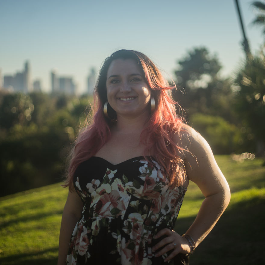 Woman with pink hair in a floral dress smiling outdoors with city skyline in the background, focusing on birthday interviews.
