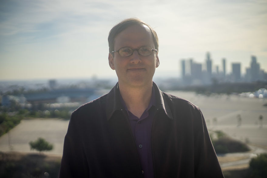 Man wearing glasses and a dark jacket standing outdoors with city skyline in background during daytime birthday interview session