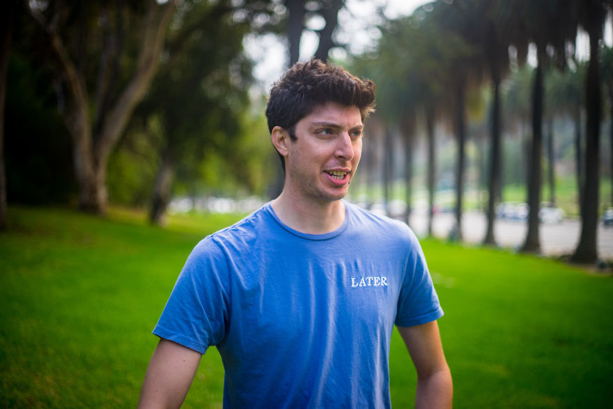 Man wearing a blue shirt outdoors, appearing engaged while interviewing people on their birthdays in a park setting