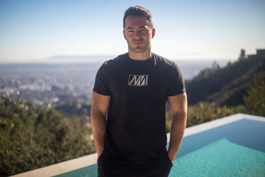 Man standing outdoors by a pool with city view in the background, focused on interviewing people on their birthdays.