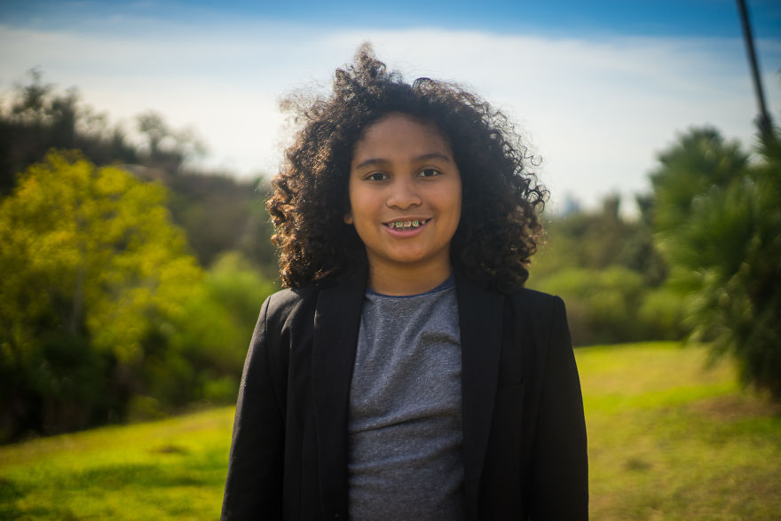 Child with curly hair smiling outdoors on birthday, dressed in a blazer and casual shirt, celebrating with nature background.