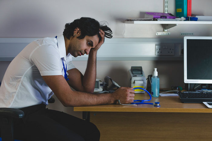 A male doctor is sitting at his office desk, he has his head in his hands and looks depressed and deep in thought. A male doctor is sitting at his office desk, he has his head in his hands and looks depressed and deep in thought.