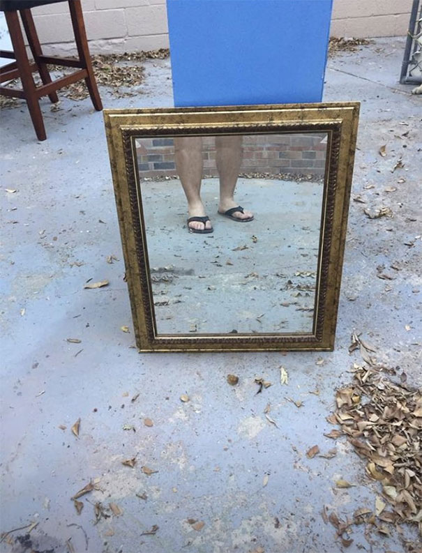 A framed mirror reflecting a person's legs in sandals, placed outside on a concrete surface.