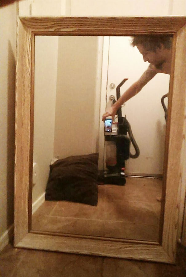 Person capturing a mirror reflection with a phone, showing a vacuum and a doorway in the background.