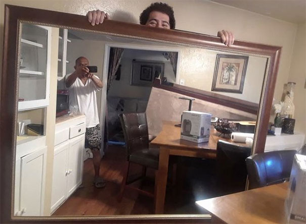 Person holding a large mirror, reflecting another person taking a photo in a dining room setting.