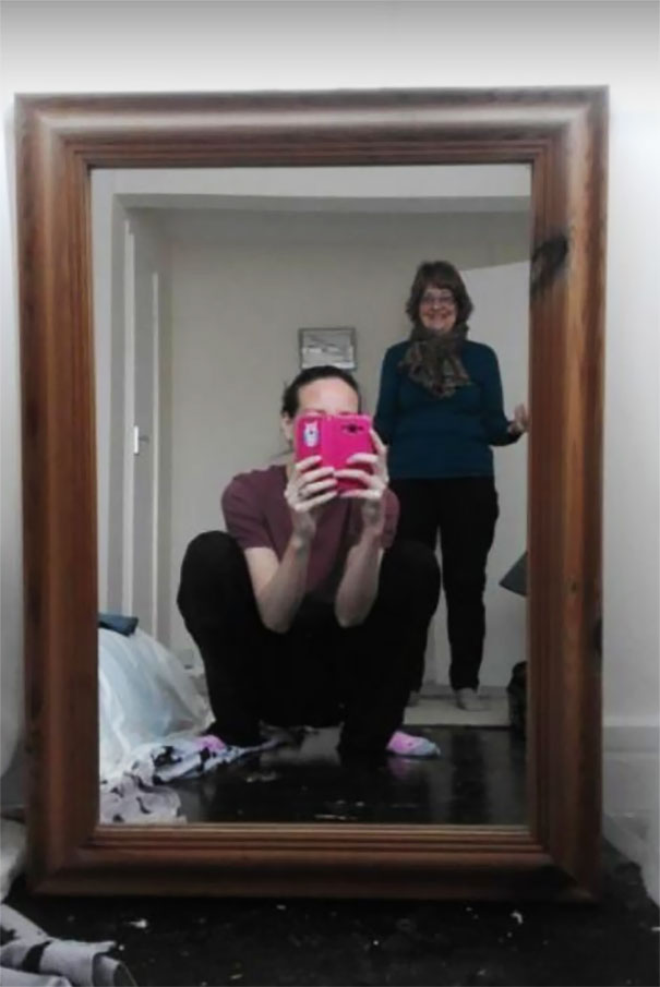 Person crouching in front of a mirror, taking a photo, with a woman smiling in the background, showcasing mirrors.