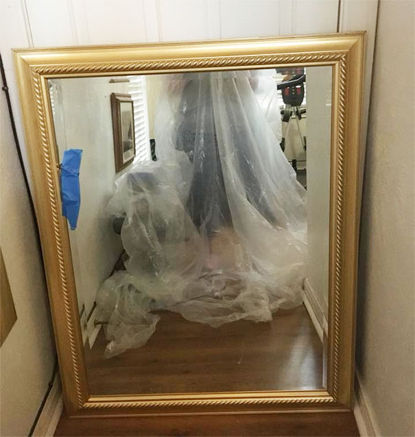 A large gold-framed mirror reflecting a person covered with plastic in a room, highlighting mirrors for sale.