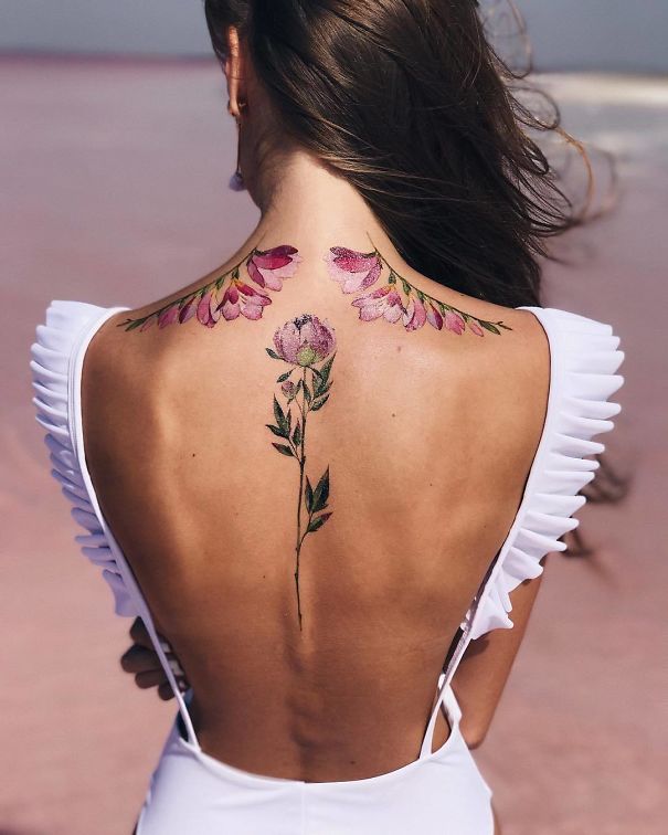 Back view of a woman with a floral spine tattoo, featuring pink flowers and greenery, wearing a white backless dress.