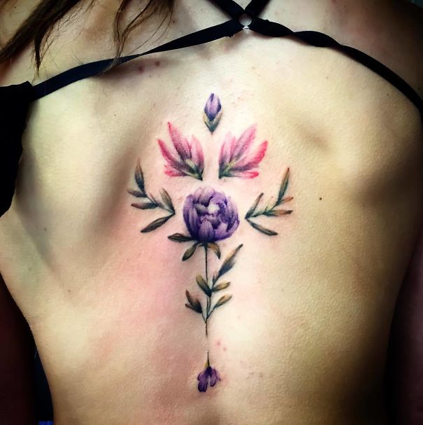 Floral spine tattoo featuring a purple flower and green leaves on a person's back.