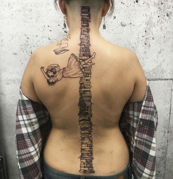 Spine tattoo featuring a stack of books with a falling girl, symbolizing creativity and imagination.