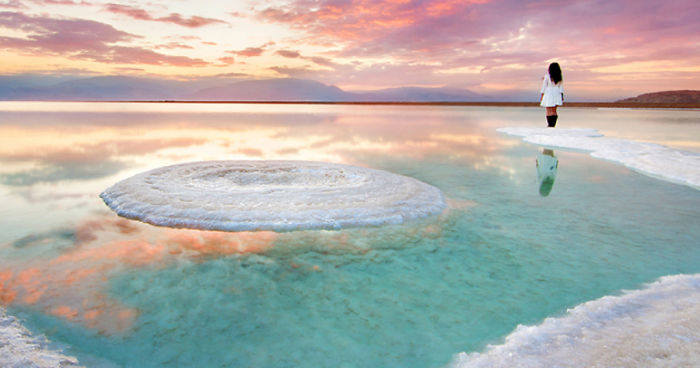 Woman standing on the edge of a clear saltwater pool surrounded by breathtaking landscapes at sunset.