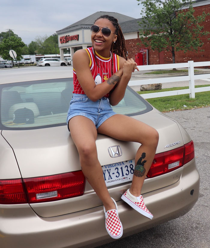 Smiling Sierra Alexis sitting on a car, showcasing a leg tattoo by a talented tattoo artist.