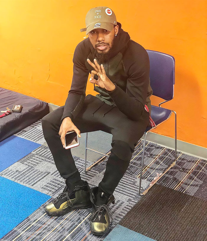 Man sitting casually on a chair, wearing a cap and holding a phone, highlighting tattoo artist skill and humor.