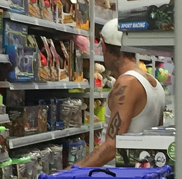 Man with a poorly done tattoo in a store aisle, wearing a white tank top and cap.