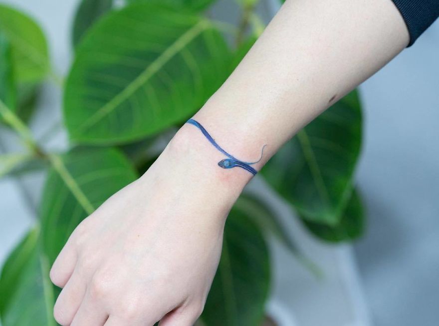 A unique snake tattoo by a Korean artist wrapping around a wrist with green leaves in the background.