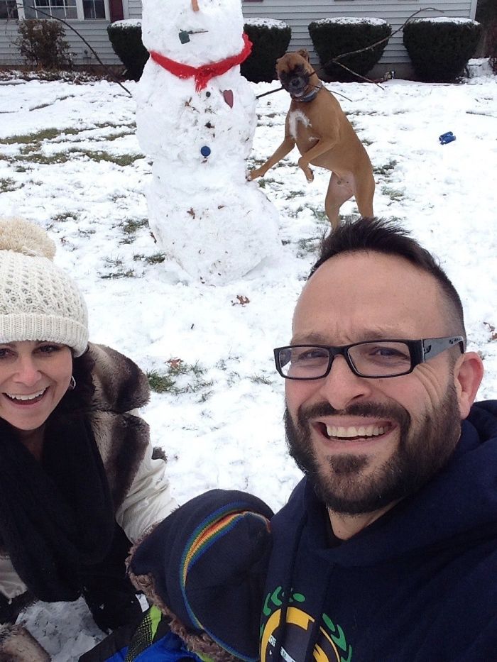 Boxer dog playfully poses with a snowman while a couple smiles for a selfie in a snowy yard.