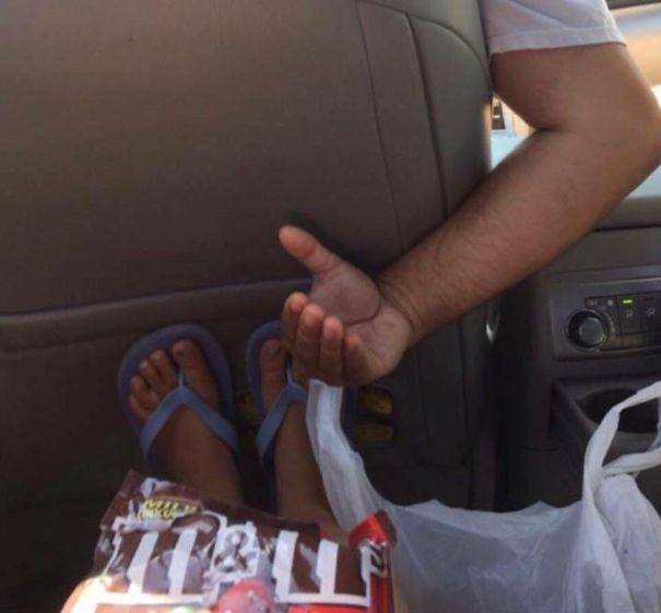 A dad humorously signaling for a snack from the backseat, while kid's feet are resting near a candy bag in a car.
