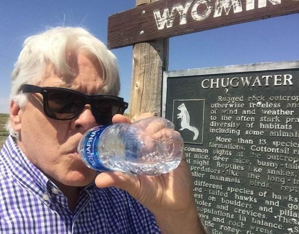A dad in sunglasses drinks water humorously in front of a Wyoming sign.