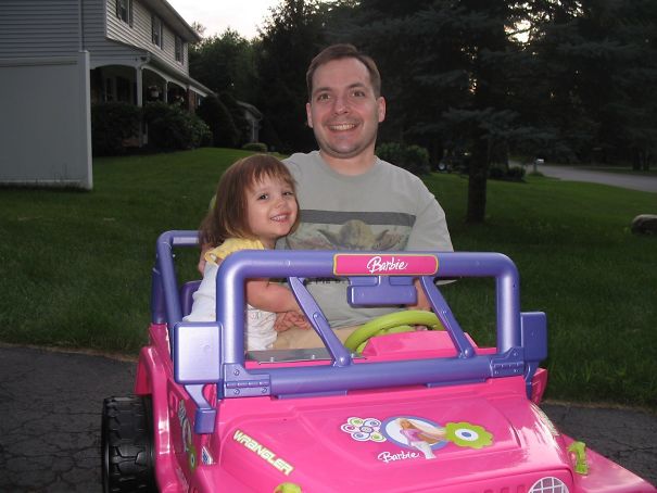 Dad and daughter in a park riding a pink toy car, smiling and enjoying a sunny day.