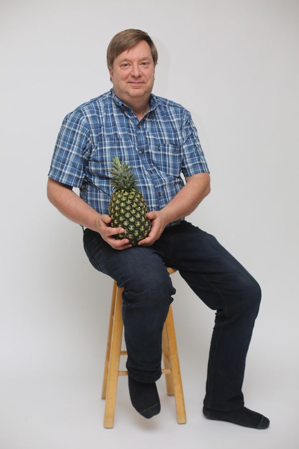 Man in plaid shirt sitting on stool, humorously holding a pineapple, representing funny dads.