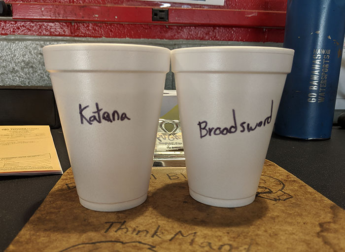 Two styrofoam cups labeled Katana and Broadsword, showcasing coworkers trolling with Asian and white stereotypes.