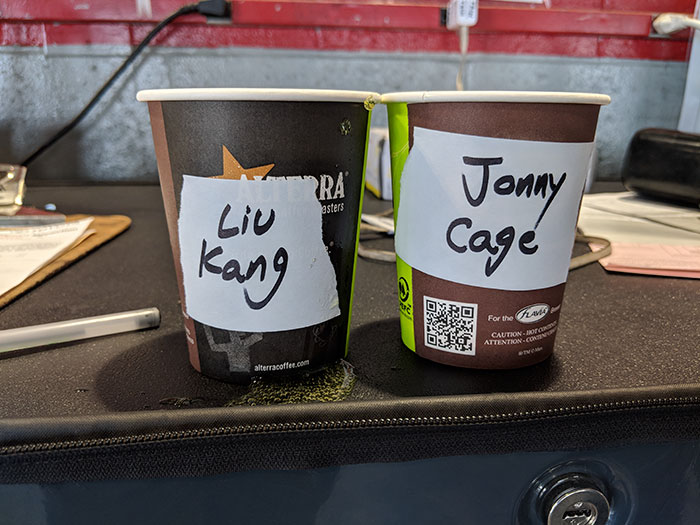 Two coffee cups on a desk labeled with Asian and white stereotype names in a humorous coworker prank setting.