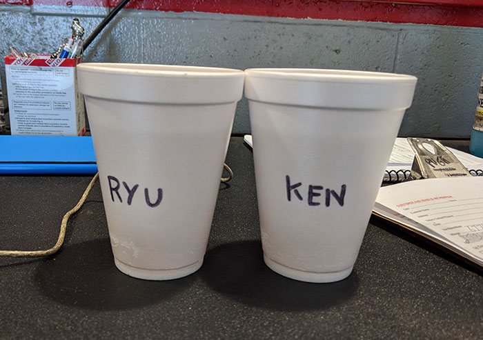Two white foam cups labeled RYU and KEN on a desk, illustrating coworkers trolling with Asian and White stereotypes.