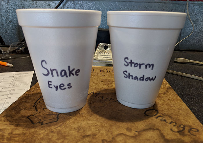 Two white foam cups labeled with nicknames on a desk, showing coworkers humorously trolling with stereotypes.