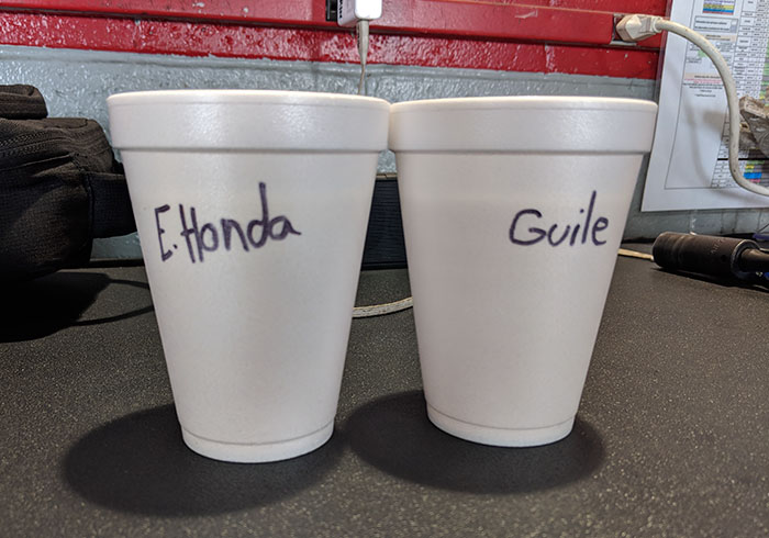 Two white foam cups with names written on them, set on a desk in a casual office environment, coworker humor.