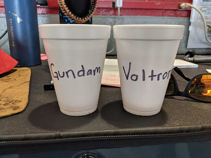 Two white foam cups labeled Gundam and Voltron, showing coworkers trolling with Asian and white stereotypes.