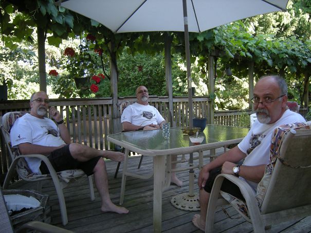 Three funny dads relax on a patio under an umbrella, each sporting glasses and white shirts, surrounded by greenery.