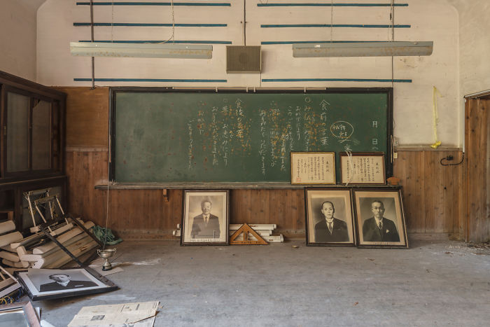 Abandoned classroom in Japan with old portraits, dusty floor, and handwritten notes on a green chalkboard.