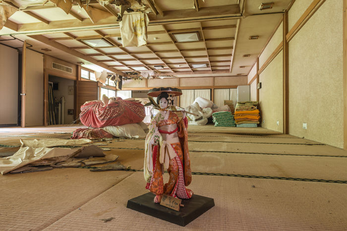 Traditional doll standing in a dusty tatami room of an abandoned place in Japan with old stacked bedding and peeling ceiling panels