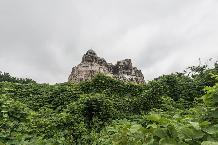 Overgrown abandoned place in Japan featuring a weathered rock formation surrounded by dense greenery under a cloudy sky.