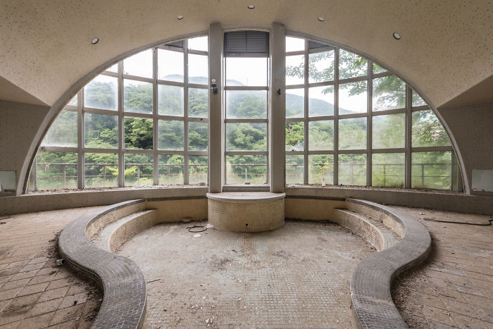 Abandoned circular indoor pool with large arched windows overlooking lush green trees in a forgotten place in Japan.