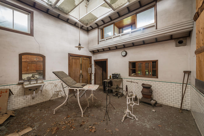 Abandoned medical room in Japan featuring old examination table, rusty stove, and peeling walls with natural light.