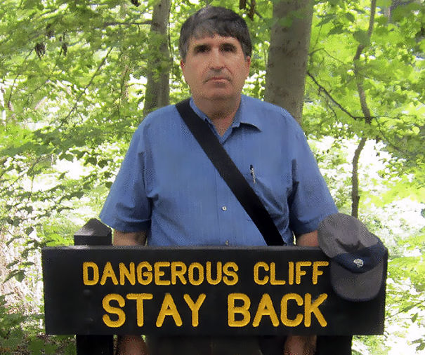 A dad humorously posing by a "Dangerous Cliff Stay Back" sign in the woods.