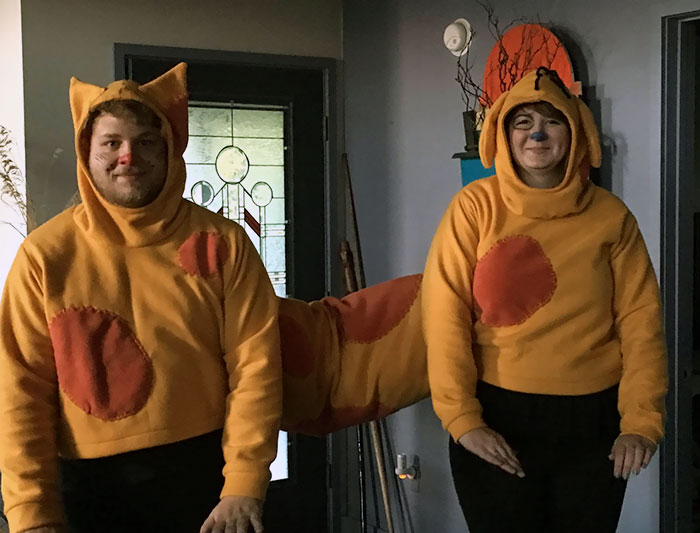 Couple in Halloween costumes, dressed as a cartoon cat and dog with connected bodies, smiling indoors.