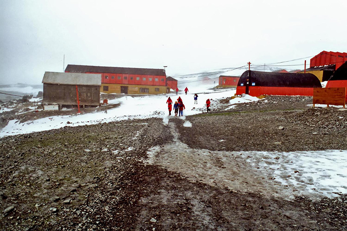Researchers in red jackets walking near snowy and rocky terrain at an Antarctica research station under cloudy skies