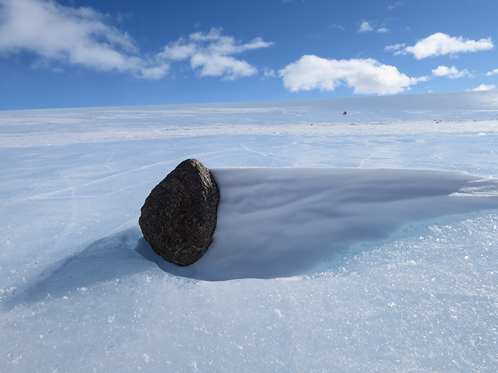 A rock partially buried in Antarctica’s icy landscape casting a long shadow under a bright blue sky.