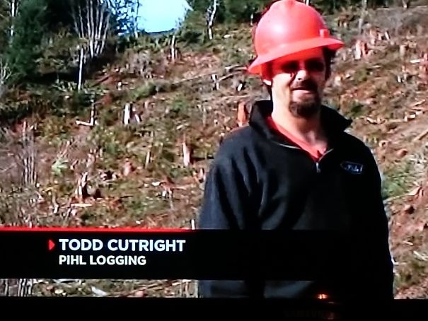 A logger named Todd stands in a clearing, wearing a red hardhat, humorously fitting the job description amidst tree stumps.